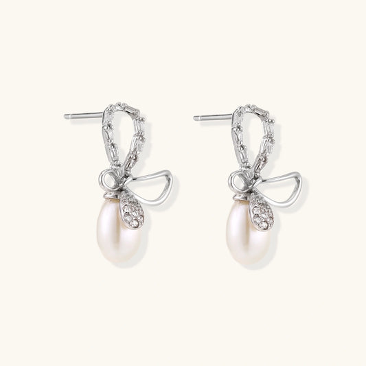 Silver bow-shaped stud earrings with baguette crystals and dangling white freshwater pearls on cream background