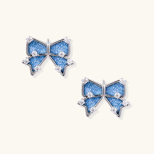 Blue denim bow stud earrings with white gold plating and crystal accents on cream background
