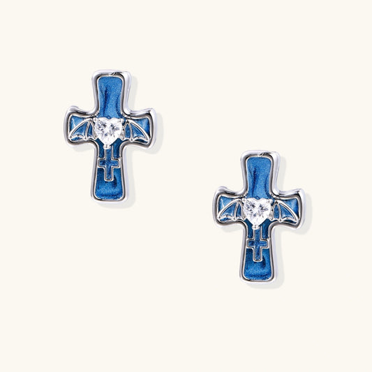 White gold-plated cross earrings with blue enamel, bat wing details, and heart-shaped zirconia center on cream background.