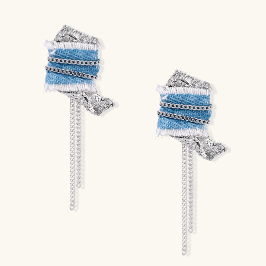 Blue denim fabric earrings with white fringe, silver chains, metallic accents, and dangling chain tassels on cream background.