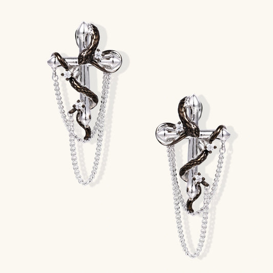 Silver dangle earrings with sculptural serpent coiled around cross shape and delicate chain accents on cream background