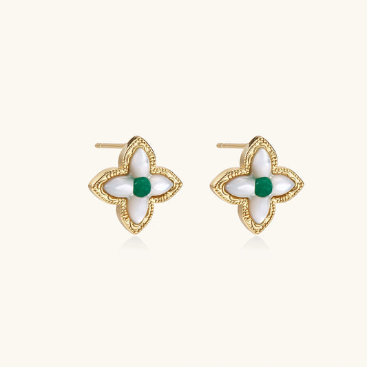 Gold quatrefoil stud earrings with white mother-of-pearl petals and emerald green gemstone center on beige background.