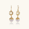 Gold sunburst charm earrings with baroque pearl drops on French hooks against cream background