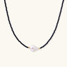 Delicate necklace with black blue sandstone beads and baroque pearl center on white background with gold accents.