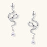 Silver snake-shaped drop earrings with crystal accents and teardrop stone dangles on cream background.
