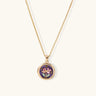 Gold-plated circular pendant with hand-painted purple enamel floral design on delicate chain against cream background